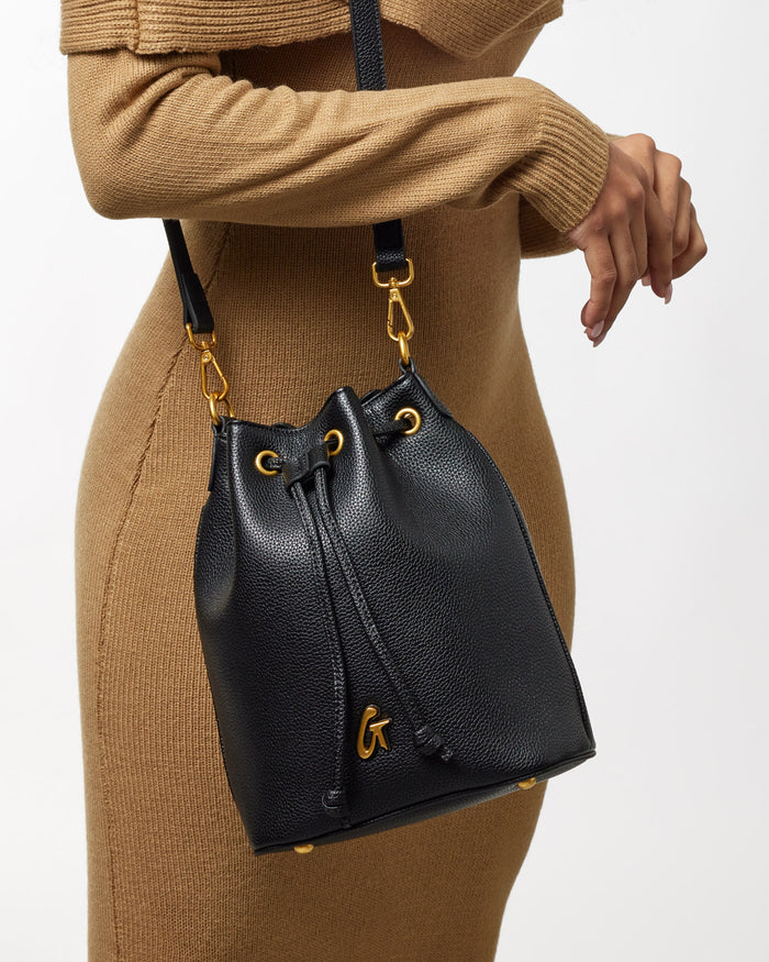 Black medium pebble bucket bag with gold hardware and a drawstring closure, carried by a person in a tan outfit.